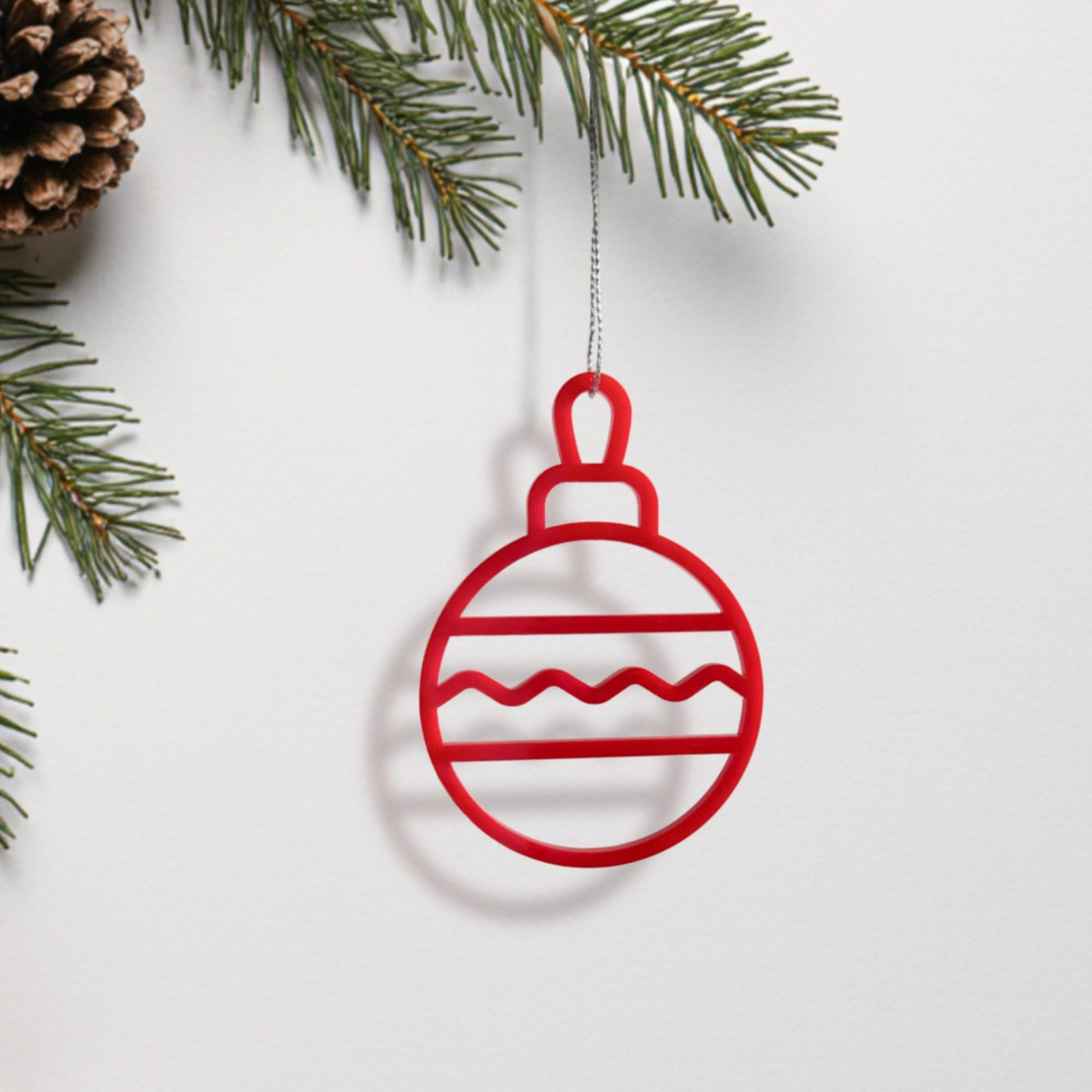 A red ornament with a wavy line design hangs from a branch of a pine tree.
