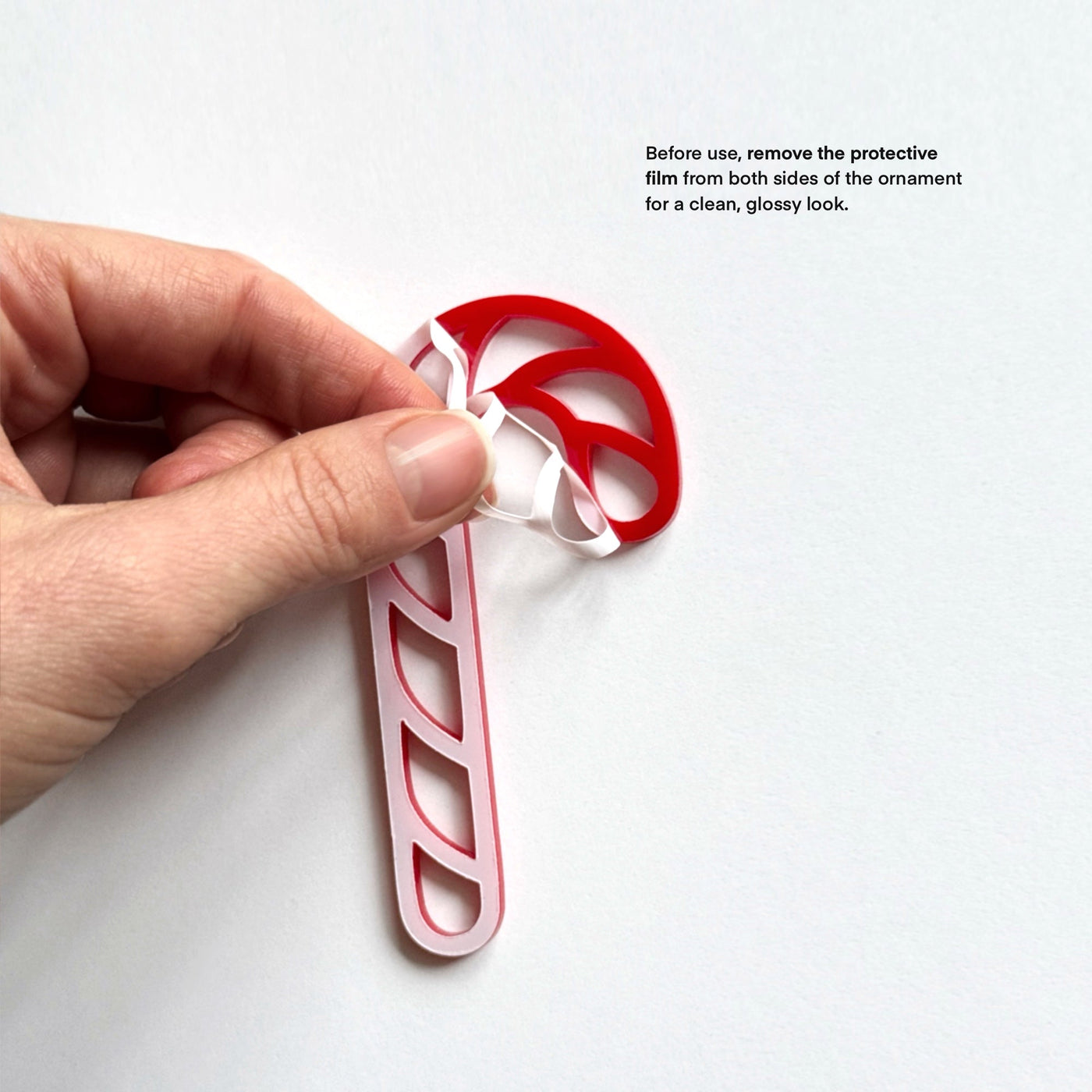 A hand is holding a red and white candy cane ornament, with instructions on how to remove the protective film from both sides of the ornament for a clean, glossy look.
