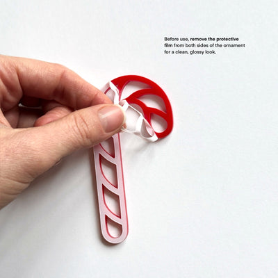 A hand is holding a red and white candy cane ornament, with instructions on how to remove the protective film from both sides of the ornament for a clean, glossy look.