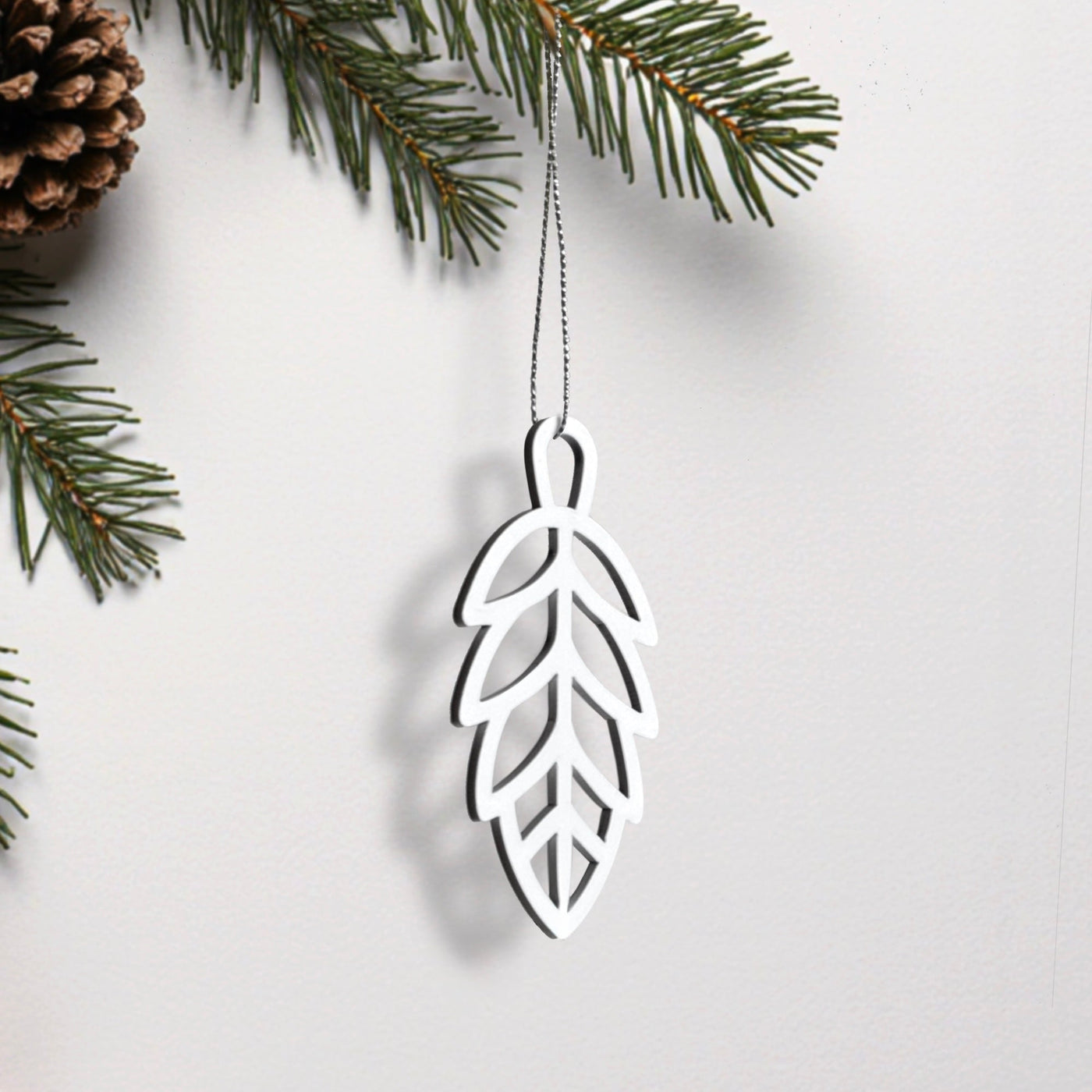 A white ornament resembling a leaf hangs from a string, with pine branches and a pine cone visible in the background.