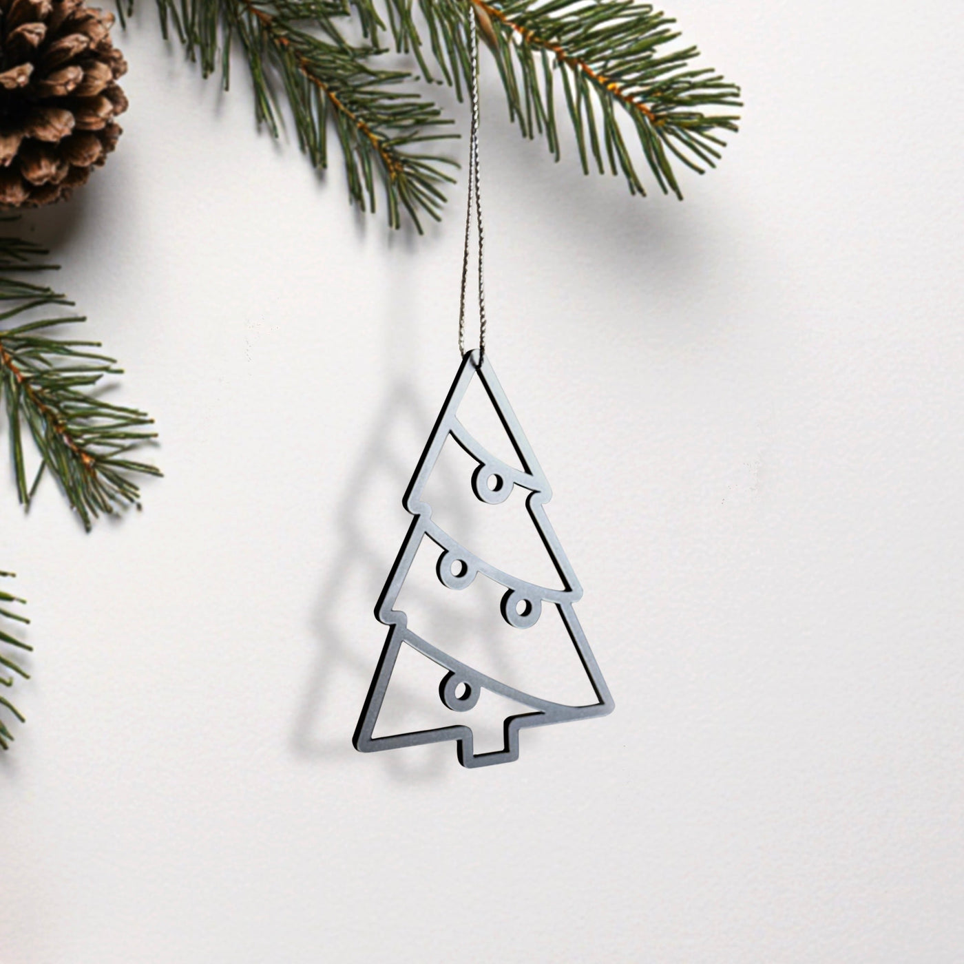 A silver Christmas ornament in the shape of a tree is hanging from a string, with pine branches and a pine cone visible in the background.