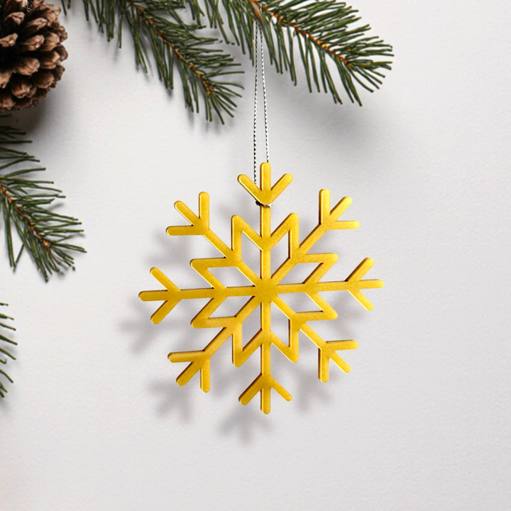 A golden snowflake ornament is hanging from a string, surrounded by pine branches and pine cones.